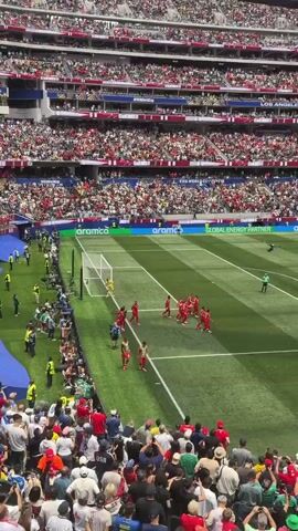 A high-angle shot shows a soccer game in progress. The field is green with white lines. Several players in red uniforms are near the goal on the right side of the frame. A goalie in yellow is in the net. Spectators fill the stands, many wearing red and white. A large screen above the stands displays text, including "SWITZERLAND" and "FIFA WORLD CUP 2026." The advertising hoardings around the field show logos, including "aramco" and "GLOBAL ENERGY PARTNER." The overall atmosphere appears to be of a lively sporting event.