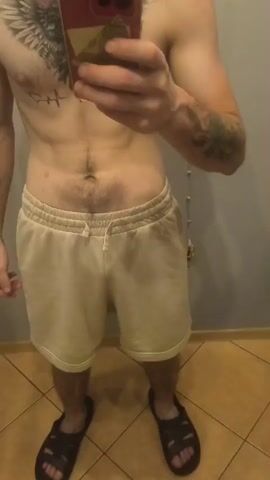 The video shows a man standing in front of a mirror, holding a phone to take a selfie. He is wearing light-colored shorts and is shirtless. The man has tattoos on his chest and arm. He flexes his arm at one point in the video. The environment appears to be a bathroom with tiled flooring and a gray wall. There is a toilet paper holder visible on the wall.