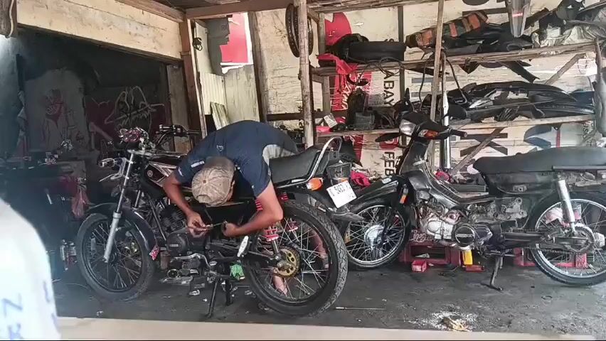 A mechanic is working on a motorcycle in a cluttered workshop. He is wearing a blue shirt, a cap, and is bent over the motorcycle, appearing to be fixing or adjusting something. There are several other motorcycles in the workshop, along with shelves filled with motorcycle parts. The workshop appears to be an indoor space with a wooden roof and walls. The lighting is dim, suggesting it might be late in the day or the workshop is not well-lit. The overall scene depicts a busy but disorganized motorcycle repair shop.