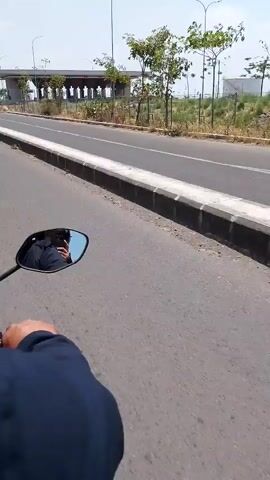 The video shows a first-person perspective from a motorcycle riding on a paved road. The rider's left arm and hand are visible in the lower left corner of the frame, along with a motorcycle mirror reflecting the rider's face and the sky. The road has a white line on the right side and a raised concrete curb. Beyond the road, there is a grassy area with scattered trees. In the background, a modern structure with arched supports is visible, along with streetlights. Further in the distance, there are industrial storage tanks. Another motorcycle passes by in the opposite direction. The sky is clear and bright, suggesting a sunny day.