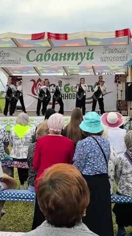 A group of young female dancers in matching black vests and white shirts perform on a stage during an outdoor event. They are positioned across the stage, some in the front and some in the back, performing synchronized arm movements and leg extensions. A banner with Cyrillic text and floral imagery is displayed behind them. In front of the stage, an audience of predominantly older individuals is seated on benches, facing the performers. The audience members are dressed in casual clothing, with some wearing hats. The setting appears to be a park or open field, with greenery visible in the background. The sky is overcast.