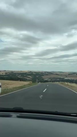 The video shows a car's dashboard and a winding asphalt road in a rural landscape under a cloudy sky. The road curves to the right, descending into a valley dotted with trees and fields. In the distance, several buildings and wind turbines are visible. The lighting suggests it is daytime.