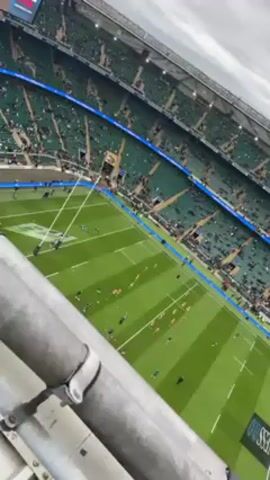 A video, taken from a high vantage point, shows a rugby match in progress in a large stadium. The field is green and well-marked with white lines. Players in different colored jerseys are scattered across the field. The stadium is mostly full of spectators. Above the stadium, the sky is overcast. A metal railing obscures the foreground of the video. Red lights flash intermittently near the upper edges of the stadium.