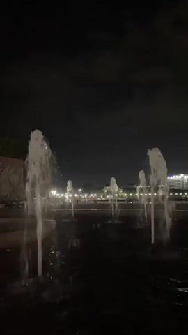 The video shows a series of fountains operating at night. The fountains are spraying water upwards in columns, and the water is illuminated by an unseen light source. The background is dark, with some faint lights visible in the distance. The camera is positioned low to the ground, looking up at the fountains. There are no people or animals visible in the video.