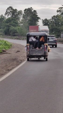 The video shows the interior of an auto-rickshaw in motion. Five people are packed inside, seated on the bench seat and in the open rear cargo area. The passengers are dressed in casual clothing. The auto-rickshaw is traveling on a paved road, with a grassy shoulder and trees lining the side. In the background, a building with a tall tower and utility poles are visible, suggesting an urban or semi-urban setting. The sky appears overcast, indicating a cloudy day. The overall mood seems to be one of practical, crowded transportation. There is no visible text in the video. The location context is Sarawadi, India.