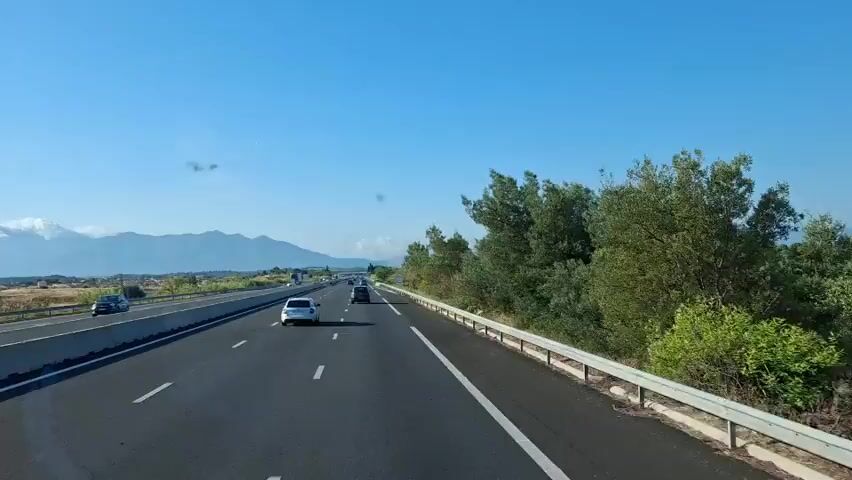 The video shows a view from a moving vehicle on a highway with a clear blue sky. Mountains are visible in the distance, partially covered by clouds. There are trees and vegetation on the right side of the highway, and a concrete barrier separates the lanes. Several cars and a truck are driving on the highway. A sign with "FM 107.7" and the VINCI logo is visible near the truck.