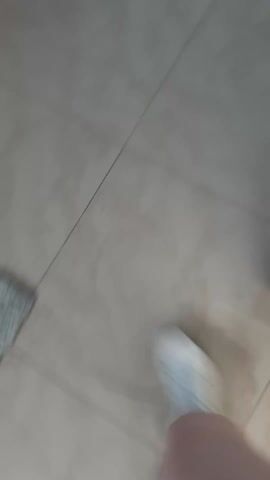 A person walks across a tiled floor. The floor is light tan or beige and appears to be made of square tiles. The person is wearing white sneakers and shorts or a short skirt. The camera is positioned looking down at the person's feet and legs as they walk.
