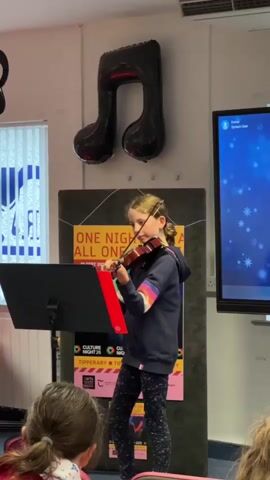 A young girl with blonde hair tied in a ponytail, wearing a dark blue hooded sweatshirt with rainbow stripes on the sleeves and patterned leggings, stands and plays the violin on a stage. A black music stand with a red sheet music folder is positioned in front of her. Behind her, a large black balloon in the shape of a musical note hangs on the wall. To the right, a large screen displays a blue, starry background. To the left, a window with vertical blinds is visible. In the foreground, the backs of several audience members are seen, suggesting an indoor performance setting. The lighting appears to be artificial, and the overall environment suggests a school or community event.