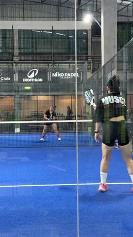 The video shows a padel tennis match played indoors. Two women are visible on the blue court. In the foreground, a woman with dark hair tied up is wearing a black cropped sweatshirt with white lettering that reads "MUSCLE FACTORY" and black shorts. She is holding a white and black padel racket and appears to be playing on the right side of the court. Her right leg is slightly forward, and her left leg is back. In the background, another woman in a black top and shorts is positioned on the left side of the court, facing the camera. She is also holding a racket. The court is enclosed by glass walls and metal fencing. Various logos, including "DECATHLON" and "Niko Padel Academy," are visible on the walls behind the court. The lighting suggests it is evening or the facility is artificially lit. The overall scene depicts an active sports environment.