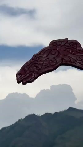 The video shows a close-up of a carved wooden horse head against a cloudy sky and mountains in the background. The horse head is dark red and intricately carved with patterns. The sky is partly cloudy with some blue visible. The mountains are covered in green vegetation and appear to be rugged. The horse head is positioned in the upper left of the frame and appears to be part of a larger structure, possibly a boat or a decorative element. The video is static, with no apparent movement.