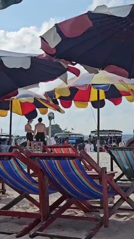 The video shows a beach scene with several colorful beach chairs and umbrellas in the foreground. In the background, there are people on the sand, some standing and some sitting. A few boats are visible in the water. The sky is blue with some clouds. A person's hand is in the lower right corner of the frame, and they are making a heart shape with their fingers. One person in the background is wearing a blue shirt with "Smile Tour" written on the back.