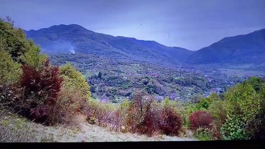 The video shows a wide shot of a valley with mountains in the background. A village is visible in the middle of the valley. The foreground is filled with trees and bushes, some of which have red and green leaves. The sky is overcast and the overall impression is of a rural and mountainous landscape. There is no indication of people or any specific actions taking place. The scene appears to be a scenic view of a valley settlement.