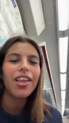 A young woman is recording a selfie video while sitting on a train. She is light-skinned with brown hair and wearing a navy blue sweater. She is talking and looking directly at the camera. Behind her are the train seats, a window, and a white ceiling with fluorescent lighting. There is also a partial view of another person sitting beside her. The video seems casually recorded with the camera focused closely on the woman's face as she speaks.