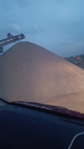 The video shows a view from inside a car. At the beginning, a very large pile of what appears to be brown sand or fine gravel fills the frame. A piece of machinery is visible on top of the pile. The view shifts as the car moves, revealing the road ahead, which is a dirt or gravel road, illuminated by the car's headlights. The sky is overcast, suggesting it is dusk or dawn.  Other vehicles are visible in the distance. The car’s interior, including the sun visor and passenger side handle, can be seen.