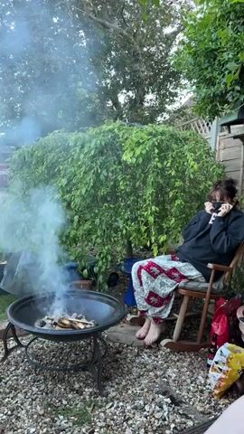 In a backyard setting, a woman sits in a rocking chair beside a smoky fire pit. She's dressed in a dark sweatshirt and patterned pants, with her face partially covered. To the left, smoke billows from the fire pit, which contains burning wood. Behind her, a weeping tree adds to the verdant backdrop. The ground is covered in gravel, and a fence is visible in the background. Bags and other items are scattered near the rocking chair.