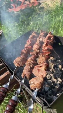 The video shows three metal skewers with meat cooking over a charcoal grill.  The skewers have twisted handles made of wood, and are arranged side-by-side over glowing charcoal.  Smoke rises from the grill, and the meat appears to be partially cooked with some browning.  The grill is set on a grassy lawn outdoors.