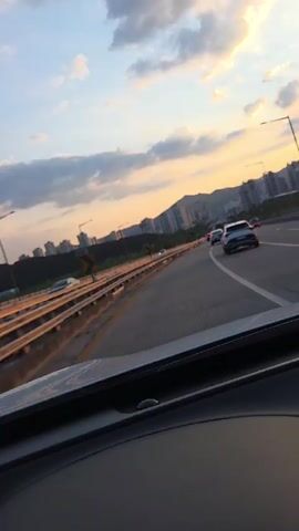 The video shows a view from the dashboard of a car driving on a highway during sunset. The sky is filled with clouds illuminated by the orange glow of the setting sun. On the right side of the highway, there are several cars driving ahead. On the left side, there is a metal barrier and then a hillside with buildings and trees. The camera is looking forward through the windshield.