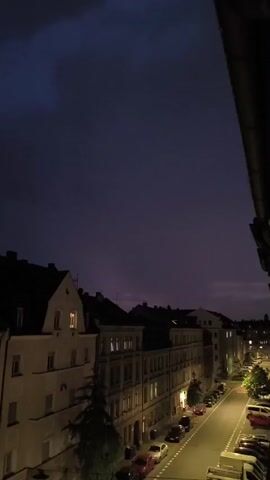 The video shows a view from a balcony overlooking a street in a European city at dusk. The sky is a deep purple and appears to be cloudy, possibly indicating an approaching storm, as there are occasional flashes of lightning in the distance. The street is lined with parked cars and buildings with illuminated windows, suggesting the evening is settling in. The architecture of the buildings appears to be traditional European style. The overall mood is calm and slightly ominous due to the darkening sky and distant lightning.