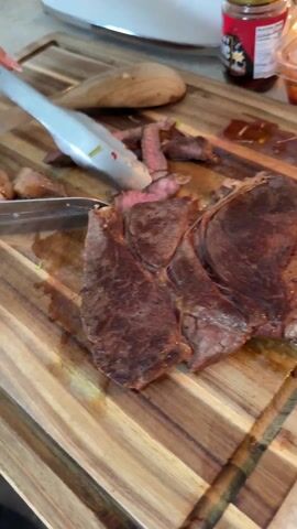 The video shows someone cutting into a large piece of cooked beef on a wooden cutting board. They use a knife in their right hand and hold the meat in place with tongs in their left. The steak is well-cooked, with a dark crust and a reddish-pink interior. The cutting board has a distinctive striped grain pattern. In the background, there are various kitchen items, including a white appliance (possibly a blender), a wooden spoon, a jar of chili oil, and a container of what looks like kimchi. The scene appears to be indoors, likely in a kitchen.
