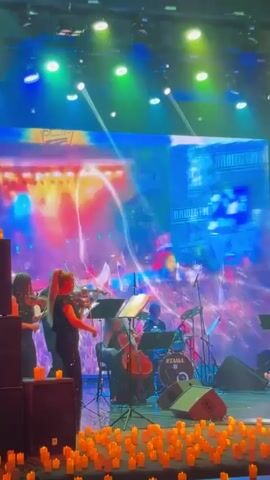 A musical ensemble performs on a stage vibrant with colorful lighting and dynamic visuals. The stage is illuminated by multiple spotlights casting bright beams, along with blue, green, purple, and pink stage lights creating a lively atmosphere. A large LED screen in the background displays shifting abstract patterns, occasionally showing distorted faces or figures.

In the foreground, three violinists, two women on the left and one woman in the center, play their instruments, reading from music stands. Behind them, a woman plays a cello, and further back, a man plays a drum kit, on which "TAMA" is visible. To the far right, another musician plays a bass guitar. Speakers are positioned on either side of the stage.

The front edge of the stage is adorned with numerous small, glowing orange objects, resembling candles or small LED lights, arranged across the entire width. The musicians are dressed in dark clothing. The performance appears to be a live concert taking place in an indoor venue at night. The location context provided is Родниковское сельское поселение, Ukraine.