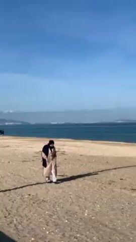 A woman is walking alone on a beach on a sunny day. The beach is mostly sand with a few small rocks. The woman is wearing a long beige coat, a black scarf, and white sneakers. She appears to be walking at a leisurely pace. In the background, there is a body of water, possibly an ocean, with land visible in the distance. The sky is mostly blue with some scattered clouds. There are a few people visible in the far distance along the water's edge. The woman's shadow is visible on the sand.
