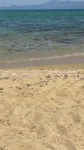 A gentle wave laps at a sandy beach. The water is clear and blue-green, with sunlight glinting off the surface. In the background, mountains rise in the distance, silhouetted against a clear blue sky. The sand is a light golden color, with small pebbles and shells scattered along the waterline. The scene is peaceful and serene, evoking a sense of calm and relaxation.