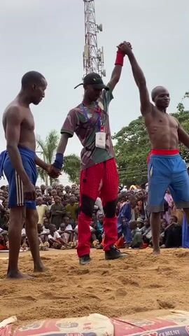 In a medium shot, two shirtless men stand on a sandy wrestling ring with the referee in the middle raising the arm of the wrestler on the right, signifying his win. Both wrestlers wear shorts, one blue with white stripes and the other blue with a red waist band. The referee wears a camouflage t-shirt, red pants, and black shoes. A large crowd sits on the ground around the wrestling ring, watching the end of the match. Behind the crowd, a tall communication tower stands. The sky is overcast. There is some debris scattered around the ring.