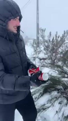 A man in a snowy landscape throws an object that appears to be a can into the air. The man is wearing a black puffer jacket, black gloves, and a black hat. The ground is covered in snow, and there are small shrubs and trees visible. The sky is overcast and foggy, with poor visibility. The man who throws the can walks towards the camera and gestures with his hands. The scene is stark due to the snow and fog.