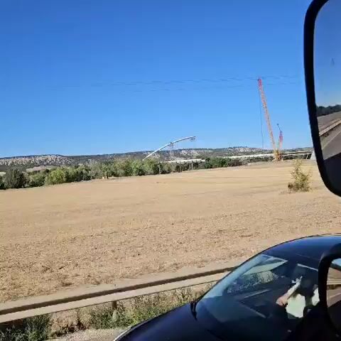 The video begins with a reflection in a truck's side mirror, showing a clear blue sky and a dry, grassy field. Two large cranes are visible in the distance, along with a long, curved bridge structure under construction. The field is mostly tan and dry, with a few scattered green bushes and trees. A power line pole stands in the field. The camera pans slightly, revealing more of the construction site and the surrounding landscape, which includes rolling hills in the background. A yellow triangular road sign with a black silhouette of a person walking is visible near the power line pole. The video continues to show the construction of the bridge, with the cranes actively working. The perspective shifts, showing the road and guardrail in the foreground as the truck moves along. The scene is bathed in bright sunlight, indicating a clear day.