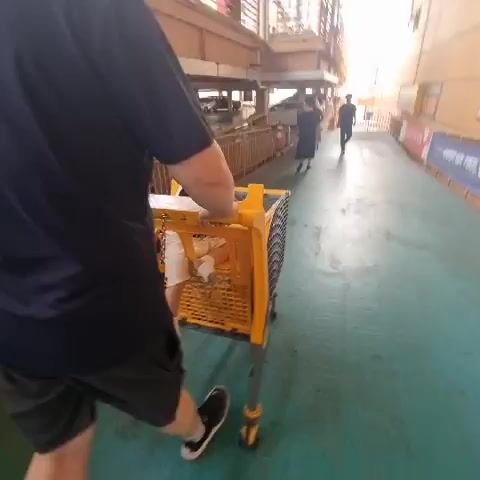 A person in a dark blue shirt and dark pants pushes a yellow shopping cart down a ramp. The shopping cart is empty. In the background, cars are parked in a multi-level parking garage. The scene appears to be indoors, with fluorescent lighting.