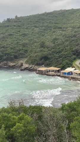 The video shows a rocky coastline with a beach, taken from an elevated position. The water is a clear turquoise color. The shore is lined with rocks and green foliage. Two small structures with roofs sit near the water's edge, one with a blue wall. A dense green forest covers the hill in the background. The sky is overcast and gray. The camera pans slightly to the left, revealing more of the coastline and the open sea. The water is choppy, with small waves visible.