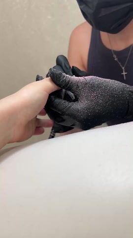 A manicurist wearing a black face mask and black gloves is using a nail drill to shape a client’s fingernail. The client’s hand rests on a white surface with pink specks scattered around. The manicurist wears a black tank top and a necklace with a cross pendant. The background is a beige wall.