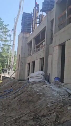 A construction site is shown on a sunny day. The unfinished building is made of concrete and light-colored blocks. A concrete pump truck is positioned to the left, with its boom extending towards the upper levels of the building. Construction workers are visible on top of the structure, engaged in some sort of building activity. There are piles of dirt and construction materials on the ground, including white bags and a blue bucket. Trees can be seen in the background.