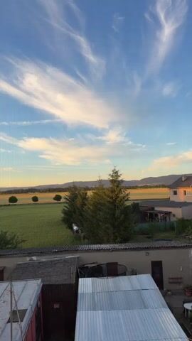 This is a vertical video shot from an elevated viewpoint, likely a balcony or upper floor window, looking out over a rural landscape at dusk. The sky is a vibrant blue with streaks of wispy white clouds illuminated by the setting sun, casting a warm, golden glow on the horizon. Rolling hills in the distance are silhouetted against the sky, with hints of darker trees and lighter fields.

In the foreground, the view is partially obstructed by the roofs of several outbuildings, suggesting a residential or agricultural setting. These structures are built with corrugated metal and weathered materials, some appearing aged. To the left, a portion of a red structure is visible.

Beyond these immediate structures, the landscape opens up to fields of green and golden crops. Several trees are scattered across the fields, including a prominent cluster of evergreen trees in the middle ground, and three lone trees standing in a row in the distance. A residential building with a red-tiled roof is visible on the right side of the frame.

The time of day is clearly late afternoon or early evening, indicated by the warm light and the long shadows that would be present. The weather appears clear and pleasant, with the beautiful sky suggesting good conditions. The overall mood is peaceful and serene, capturing the quiet beauty of the countryside as day transitions into night. There is no visible text in the video.