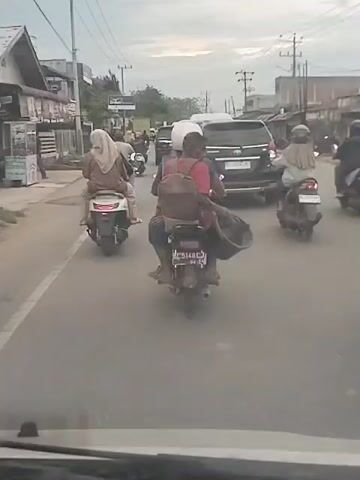 The video shows a view from inside a car driving on a street with moderate traffic. In front, several motorcyclists are also driving. One motorcycle carries two passengers: the driver wearing a white helmet and a passenger with a backpack, holding a large black tub. The motorcycle's license plate, BE 5148 CU, is partially visible. The other motorcyclists are wearing headscarves. A black SUV is driving close behind the motorcycle. Buildings and utility poles line the sides of the road, and the sky is overcast.