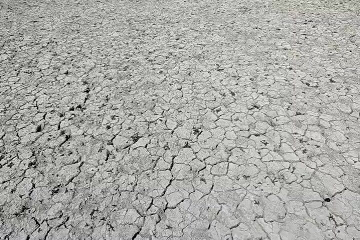 A drone flies low over a cracked, dry earth surface. The camera then pans up to reveal a distant castle-like structure situated on a hill or island. The sky is blue with a few clouds, and there is a line of green vegetation between the dry ground and the structure.