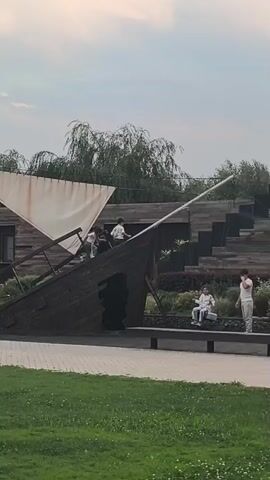 The video shows a play structure resembling a ship's hull and mast, located in an outdoor park-like setting. The structure is made of dark wood and has a large white sail attached to the mast. Several people are interacting with the structure. Two young children are climbing on the ship's hull, appearing to be playing. To the right of the ship, two adults are seated on a bench. One adult is wearing a light-colored hooded sweatshirt and appears to be looking towards the children, while the other adult, wearing a light-colored t-shirt and pants, is standing and gesturing, possibly talking to the seated person. In the background, there are trees, including a weeping willow, and a modern building with wooden siding. The sky is overcast, suggesting cloudy weather. The scene takes place during daylight, likely late afternoon or early evening, given the soft lighting. The overall atmosphere appears relaxed and recreational. The video does not contain any visible text. The location is Komsomolskoe, Kyrgyzstan.