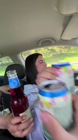 The video begins inside a car, showing a few hands holding cans and a glass bottle, as well as a person driving. A cheer occurs and the camera angle shifts to a view from the front windshield as the car moves down a road lined with trees on either side. The sky is blue with scattered white clouds. A can is held up near the dashboard.
