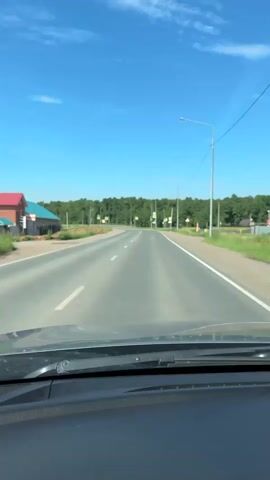 The video shows a view from the driver's seat of a car traveling on a paved road. The road is two lanes wide with white dashed lines marking the lanes. The sky is clear and blue with a few wispy white clouds. A line of green trees runs along the right side of the road. There are several traffic signs and streetlights visible along the roadside, including signs indicating pedestrian crossings and traffic lights. On the left side of the road, there is a building with a blue roof and red walls, and some construction materials. The car is moving forward at a steady pace.