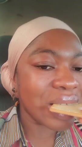A person with brown skin, wearing a peach-colored head covering and a striped shirt, is eating something that appears to be a cracker. They look slightly up while chewing. The setting seems to be inside a car, given the interior background and the person's proximity to the camera.