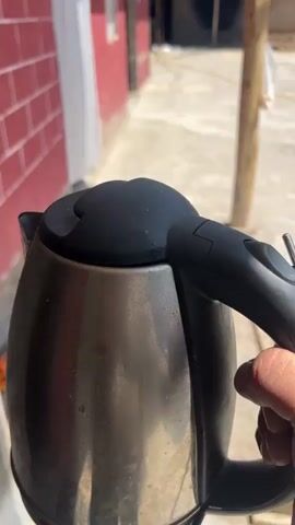 A person's hand is holding a stainless steel electric kettle. The kettle has a black lid and handle. The background is out of focus and shows a red brick wall, some white fabric, and a wooden structure. The lighting suggests it is daytime and sunny.