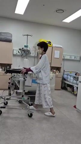 A person wearing a light-colored, patterned hospital gown and pajamas, along with a light-colored face mask, is standing and holding onto a walker in what appears to be a hospital room. The walker is a metal frame with wheels. The person's hands are resting on the grips of the walker. Behind the person, a hospital bed is visible, along with medical equipment and cabinets. The lighting in the room is from overhead fluorescent lights. The person's facial expression is not clearly discernible due to the mask. The person appears to be in the process of walking or preparing to walk.