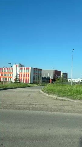 The video pans to the right showing a clear blue sky over a suburban landscape. In the foreground, a cracked asphalt road is visible. Beyond the road is a strip of green grass and then a modern, multi-story building with a white, grey, and orange facade. To the right of this building, there are several playgrounds with colorful equipment and other structures. The video continues to pan right, revealing more of the playgrounds and some smaller, low-rise buildings in the distance. The overall impression is of a bright, sunny day in a developed area that includes recreational facilities.