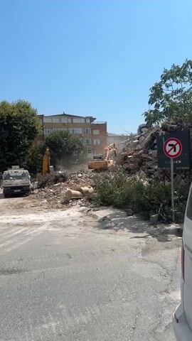 A construction site with two excavators operating near a pile of rubble under a clear blue sky. In the background, a multi-story building is visible. A white truck is parked on the left side of the frame. A signpost with a "no left turn" symbol and a logo is placed on the right. The ground is a mix of asphalt and dirt.