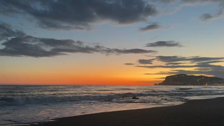 The video shows a sunset over the ocean. The sky is a mix of orange, yellow, blue, and gray, with clouds scattered across the sky. The ocean is calm with small waves gently rolling onto the sandy beach. In the distance, there is a silhouette of a landmass with lights visible along the shore.  The beach is dark and quiet. There are no people visible in the scene.