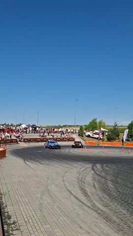 Two race cars are drifting around a paved outdoor track. The car in the foreground is blue and white with a colorful design. The car in the background is yellow. Spectators are visible on a raised platform to the left of the track. Several orange cones are placed on the track. The sky is clear and blue.