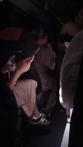 The video shows an interior view of a bus or train compartment. The lighting is dim. In the foreground, a person's arm and hand are visible, holding a mobile phone. The person is wearing a light-colored garment. Behind them, a person wearing a grey or purple jumpsuit is seated. This person is looking to their left and appears to be holding something. Further back, another person is partially visible, possibly standing or leaning. The seats are dark, and the floor appears to be carpeted in blue. A water bottle is visible on a tray table. The overall impression is of a crowded, dimly lit transit vehicle.