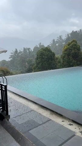 The video shows a scenic infinity pool being heavily rained on. The pool is a turquoise color, and the dark grey pool deck is also wet. In the background, there is a misty, mountainous landscape covered in lush green trees and palm trees. The sky is overcast and grey. The heavy rainfall creates ripples on the surface of the pool. There are no people visible.