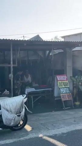 The video shows a street-side establishment with a corrugated metal roof supported by wooden beams. Two individuals are visible inside the shaded area. One person, wearing a patterned garment, appears to be interacting with another person who is wearing a white tank top. A table is set up in front of them, with what seems to be produce or food items displayed on it. To the right of the establishment, a sign is propped up, listing "WARKOP", "JUS BUAH", "PENTOL", and "FREE WIFI". A motorcycle is parked in the foreground to the left, with a large white bag secured over its rear. The environment appears to be an outdoor setting, possibly a street in a tropical or warm climate, with sunlight illuminating the scene. The overall impression is that of a small food and drink vendor.