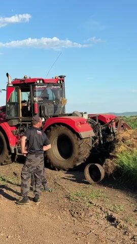 The video shows a large red tractor with its rear wheels stuck in mud and vegetation. Three men are standing around the tractor, observing the situation. One man, wearing a black shirt with a logo and camouflage pants, appears to be directing or assessing the problem. Another man, in a white t-shirt and dark shorts, stands next to him, also looking at the tractor. A third man, in a light-colored t-shirt and dark shorts, is further to the left. The tractor has a large, articulated body and appears to be partially submerged in the embankment. The ground is a dirt track, and the surrounding area is covered in green grass and vegetation. The sky is blue with some scattered clouds. A detached, circular metal object, possibly a piece of machinery, is visible near the tractor's rear wheel.